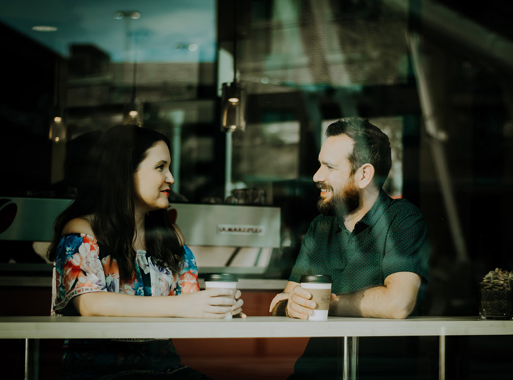 From Renew Hope and Healing: Two people sit at a café table indoors, holding coffee cups and facing each other while engaging in open communication in your relationship.