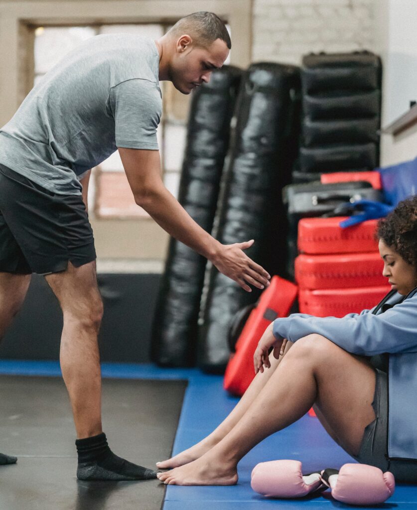 From Renew Hope and Healing: A man offers a hand to a seated woman with boxing gloves at her feet in a gym, demystifying how to reach out for support amid training mats and equipment in the background.