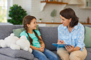 From Renew Hope and Healing: A young girl sits on a couch next to a woman holding a clipboard, both smiling and talking in a home setting—a warm moment of child counseling, with a stuffed animal beside the girl.