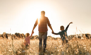 From Renew Hope and Healing: An adult holding hands with two children, walking through a sunlit field of tall grass with the sun low in the sky, capturing the warmth of love and deep relationships.