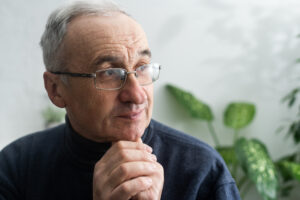 From Renew Hope and Healing: Senior man with glasses and gray hair, wearing a dark turtleneck, looking thoughtfully to the side with hands clasped under his chin; green plants in the blurred background evoke a sense of personal growth.