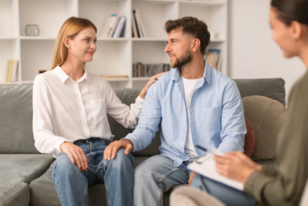 A couple sits together on a couch, holding hands and looking at each other, while a therapist with a clipboard sits across from them in an office setting from Renew Hope and Healing