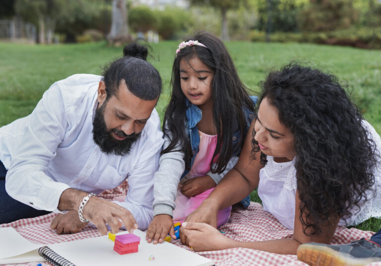 A man, woman, and young girl sit on a picnic blanket in a park, building with colorful blocks and paper on a sketchbook from Renew Hope and Healing