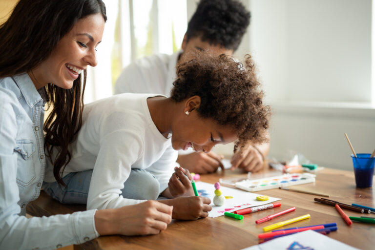 A woman and a child sit at a table drawing and coloring with markers and colored pencils, smiling and engaged in a creative activity from Renew Hope and Healing