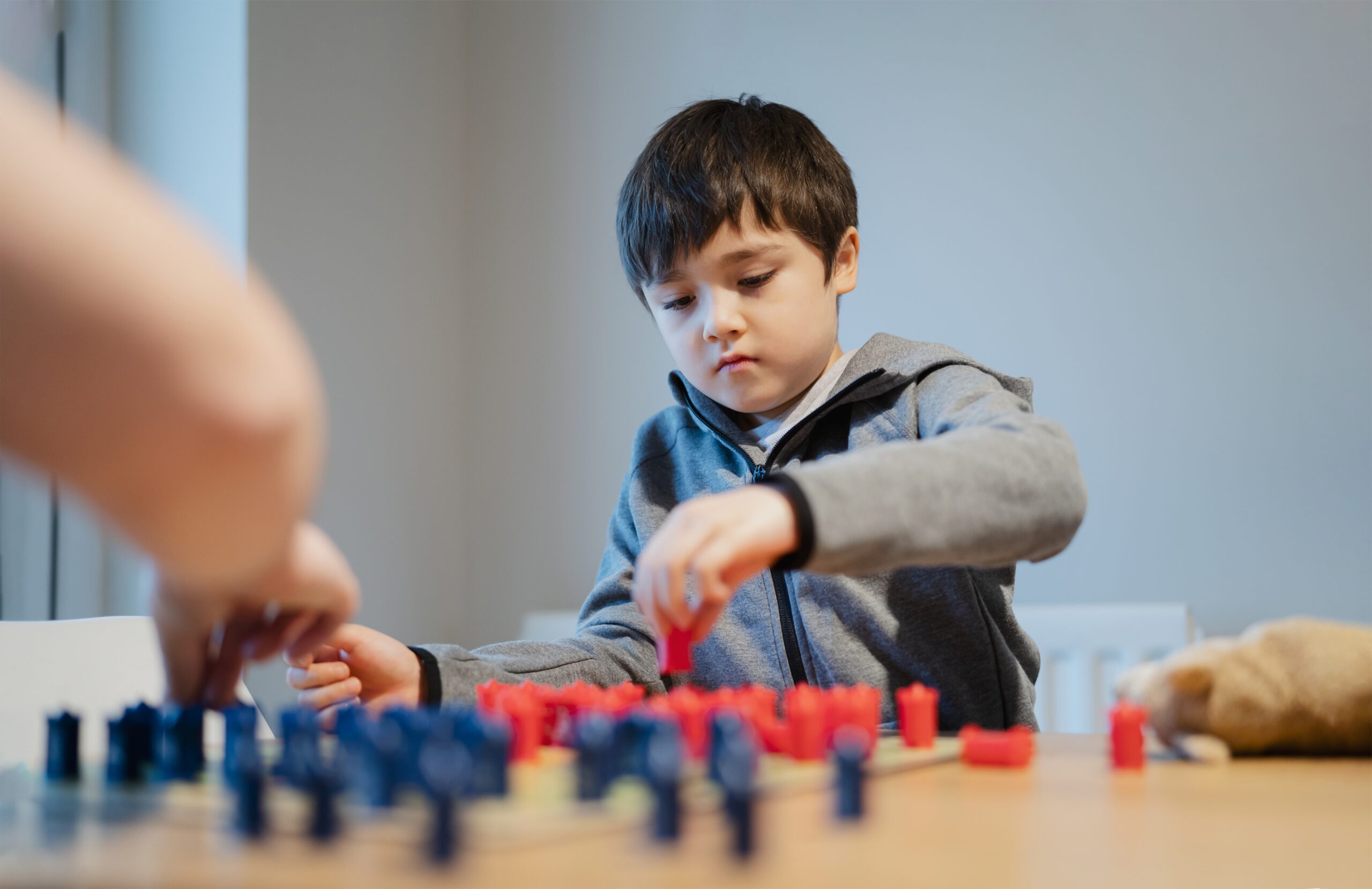 A young boy in a gray hoodie concentrates while playing a board game with red and blue pieces at a table from Renew Hope and Healing