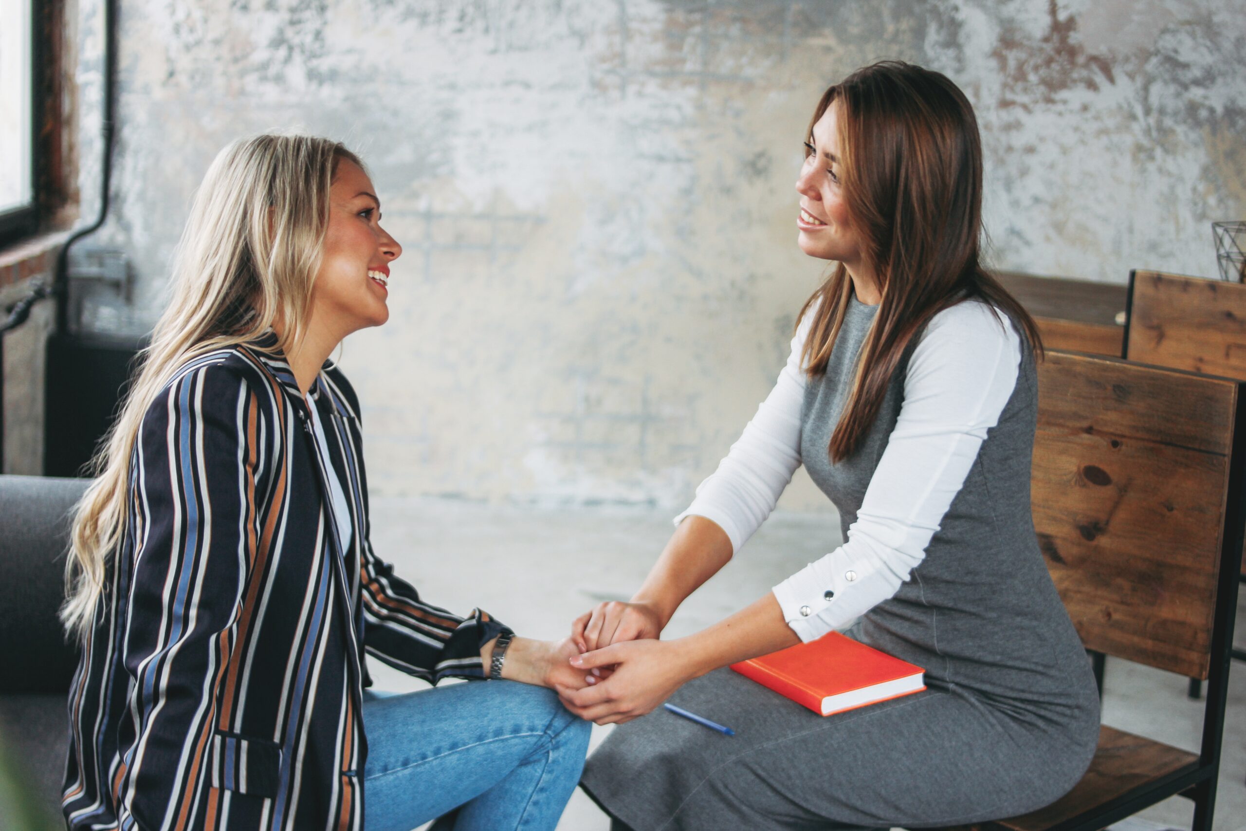 Two women sit facing each other in a casual setting, holding hands and smiling. One has a notebook on her lap from Renew Hope and Healing