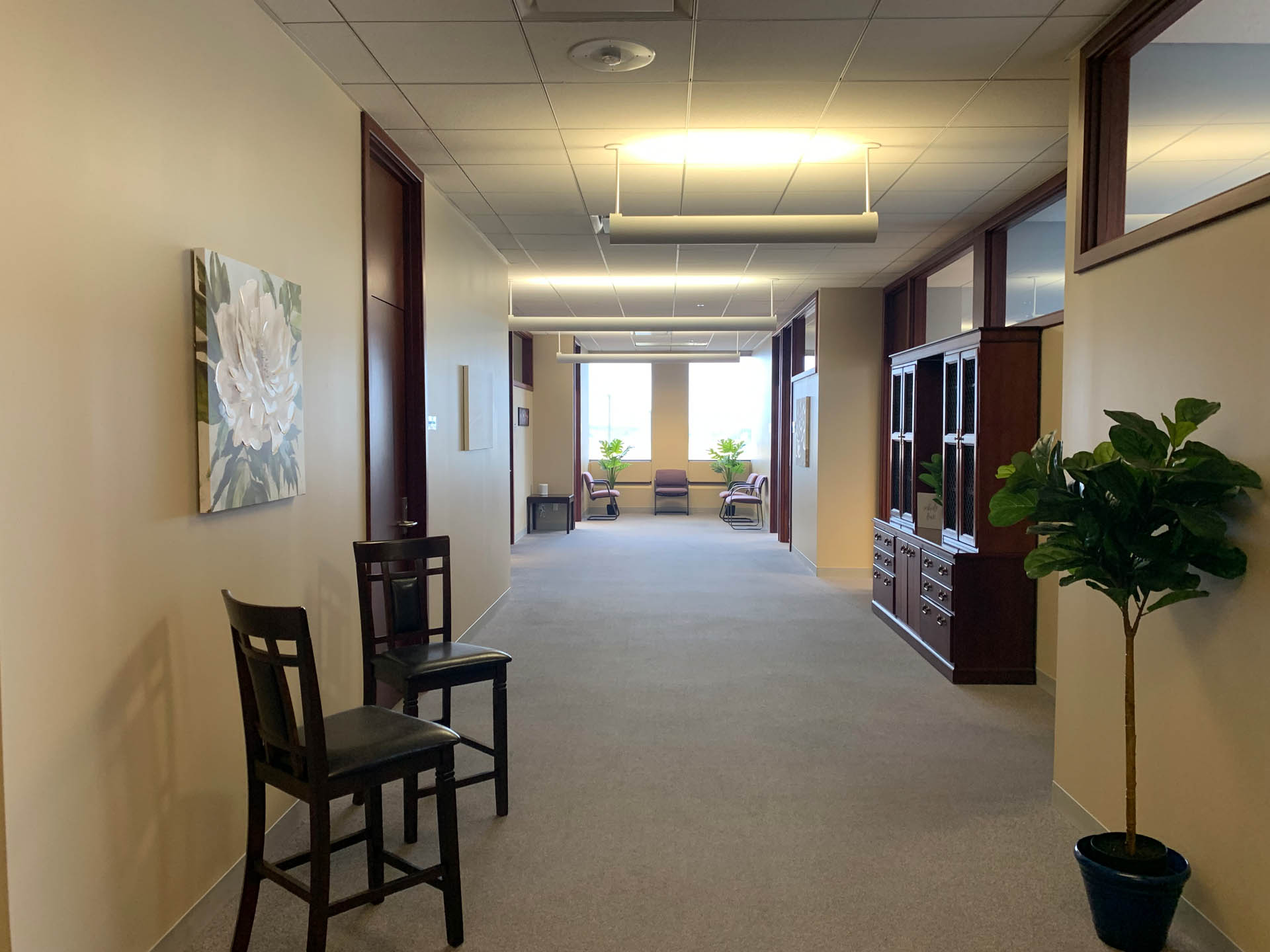 From Renew Hope and Healing: A hallway with beige walls, two chairs, a potted plant, framed artwork, and lounge seating near large windows at the end.