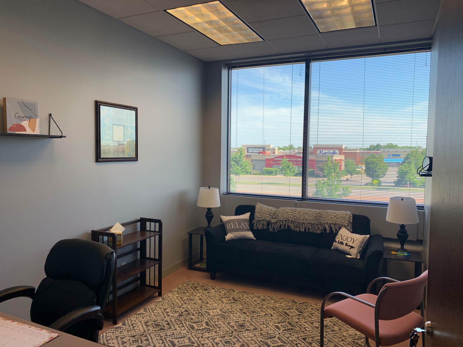 From Renew Hope and Healing: Small office with a desk, chair, couch, two lamps, shelf, and window showing a view of shops and trees outside. Pillows on couch read “Relax” and “Enjoy.”.