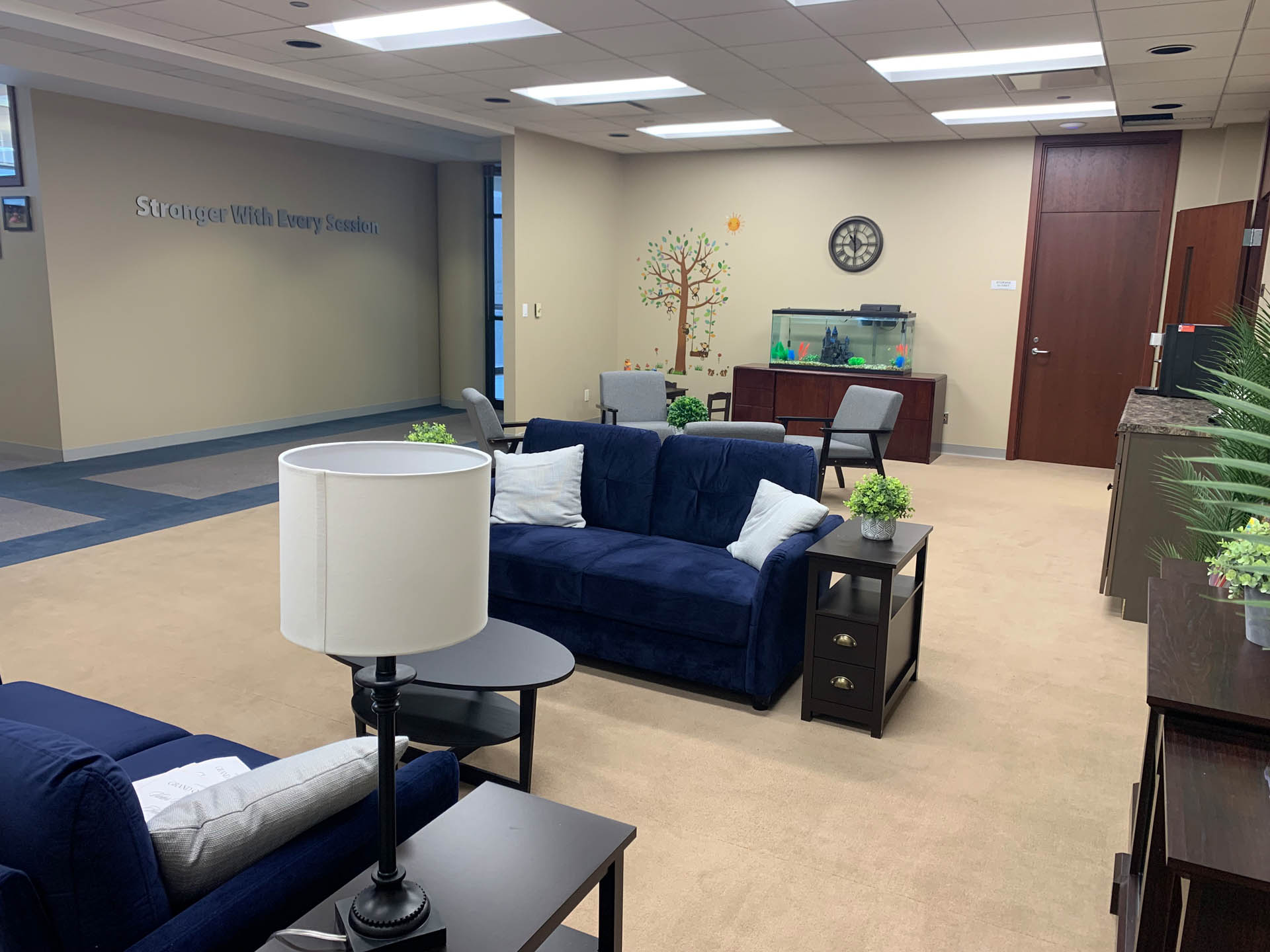 From Renew Hope and Healing: A waiting area with blue sofas, side tables, potted plants, wall decor, a fish tank, and a clock above a wooden cabinet.