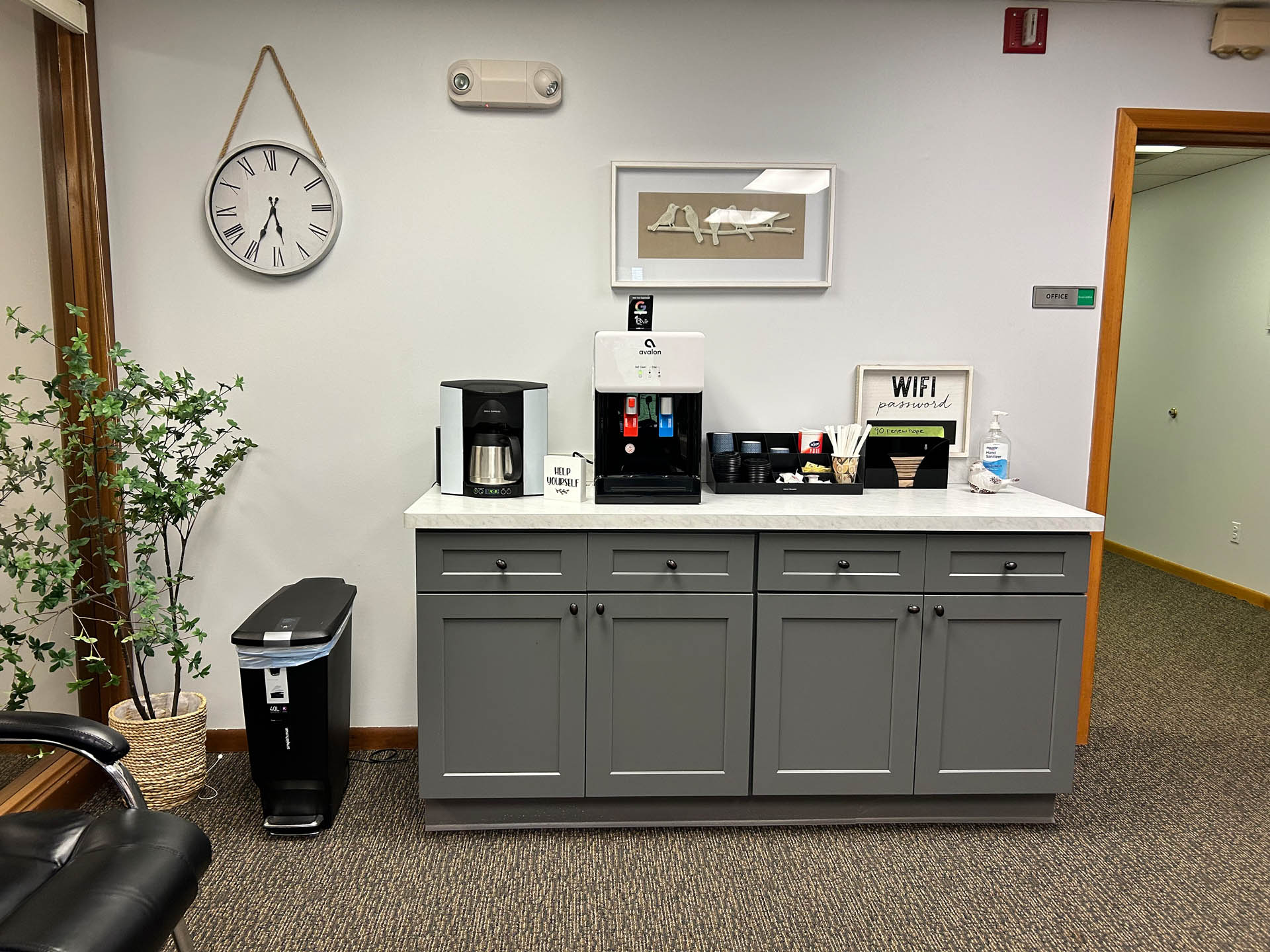 From Renew Hope and Healing: A coffee station with a coffee maker, cups, condiments, and a water bottle on a gray cabinet beneath a wall clock and framed art in an office setting.