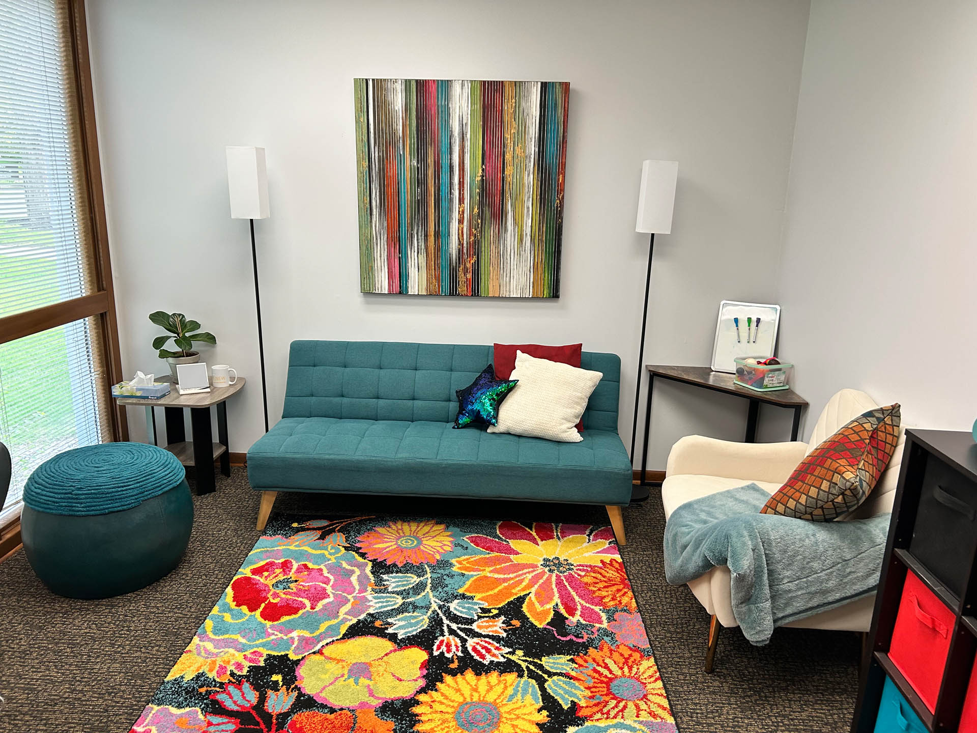 From Renew Hope and Healing: A modern, brightly decorated room with a teal sofa, white armchair, colorful floral rug, striped wall art, and floor lamps. A round pouf and small table are also present.