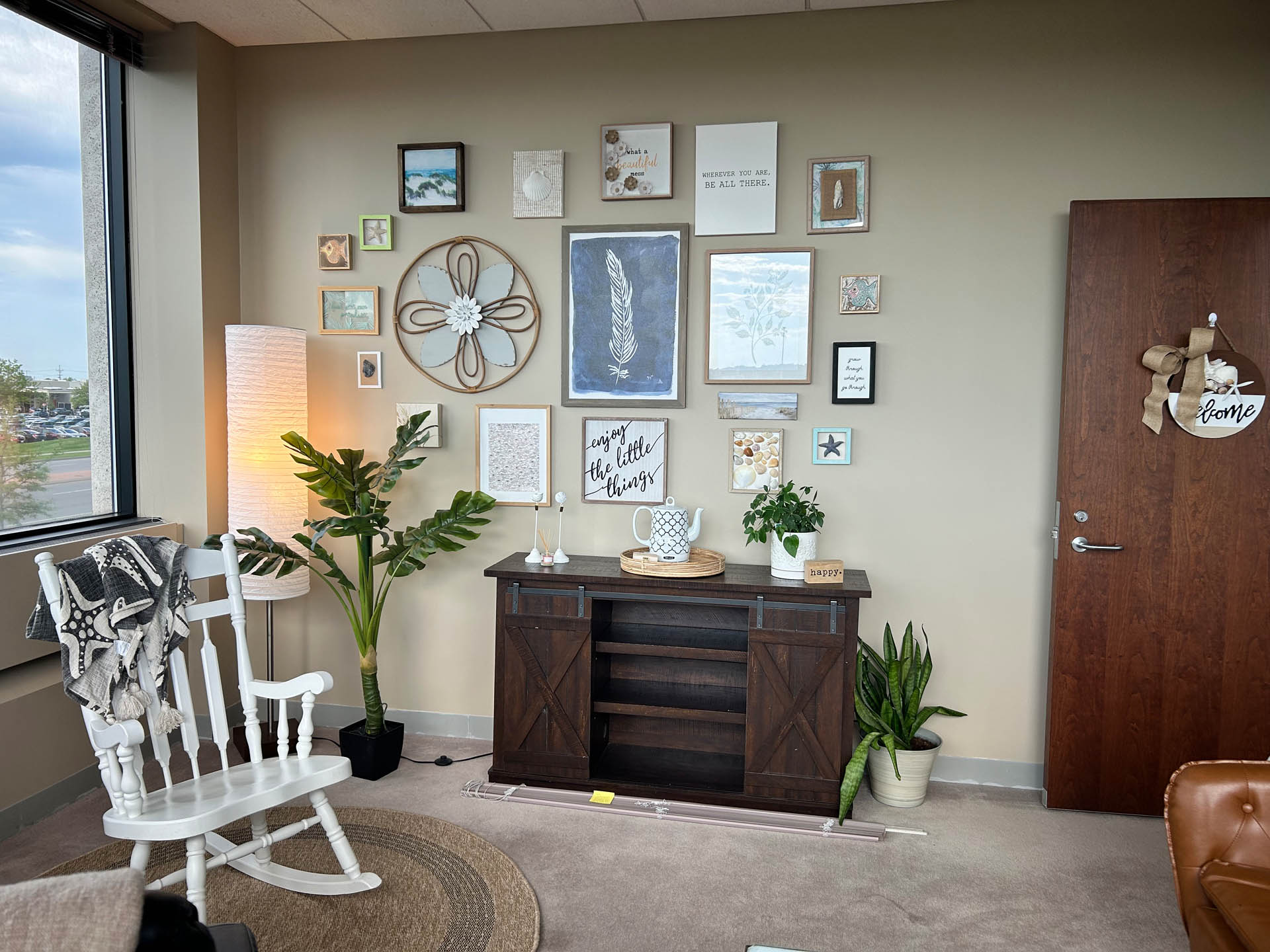 From Renew Hope and Healing: A cozy office space with a white rocking chair, floor lamp, cabinet, plants, and a wall decorated with various framed art and quotes.