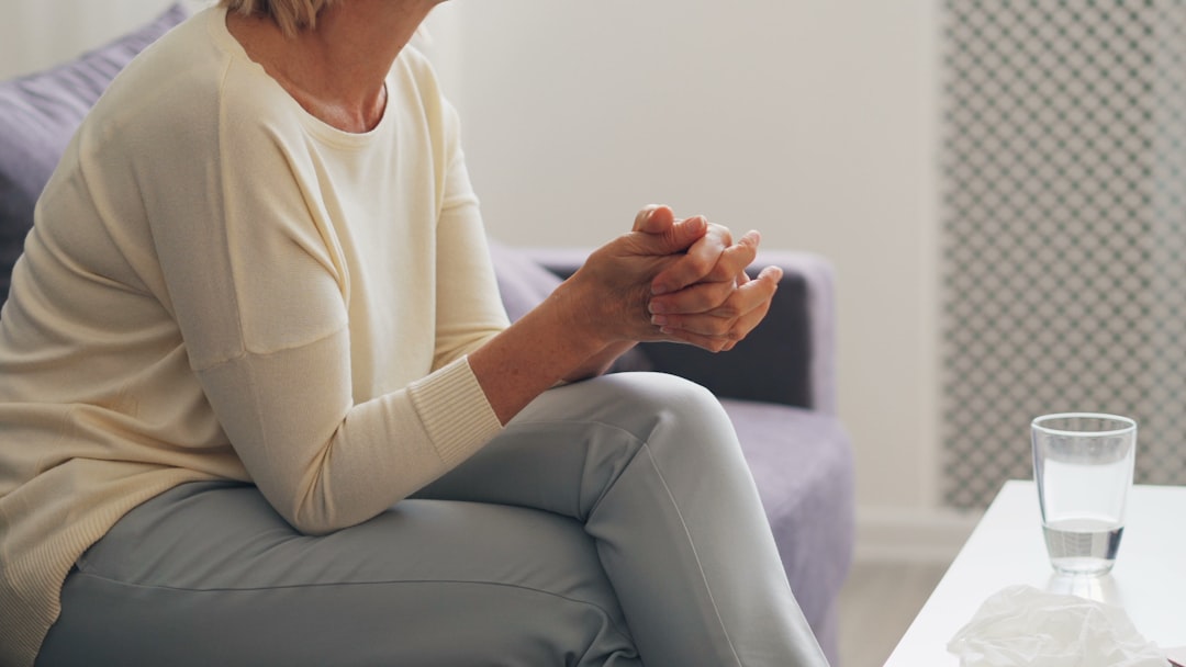 From Renew Hope and Healing: An adult sits on a couch with hands clasped, legs crossed, and a glass of water and tissue on a table nearby, suggesting a calm moment during adult therapy.