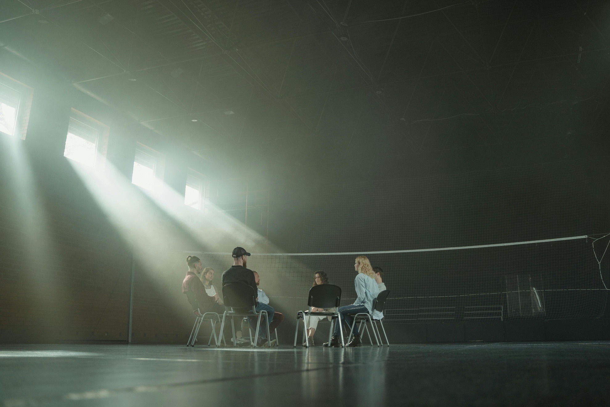 From Renew Hope and Healing: A group of people sit in a circle on chairs in a large, dimly lit gymnasium, sunlight streaming through high windows during an adult therapy session.