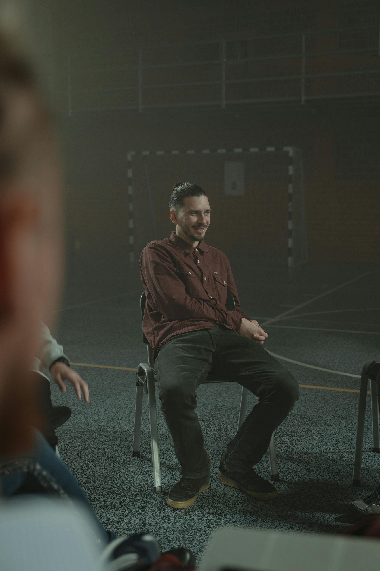From Renew Hope and Healing: A man sits on a chair in a gymnasium, smiling and leaning slightly forward, with others seated nearby, suggesting an Adult Therapy group meeting or discussion.
