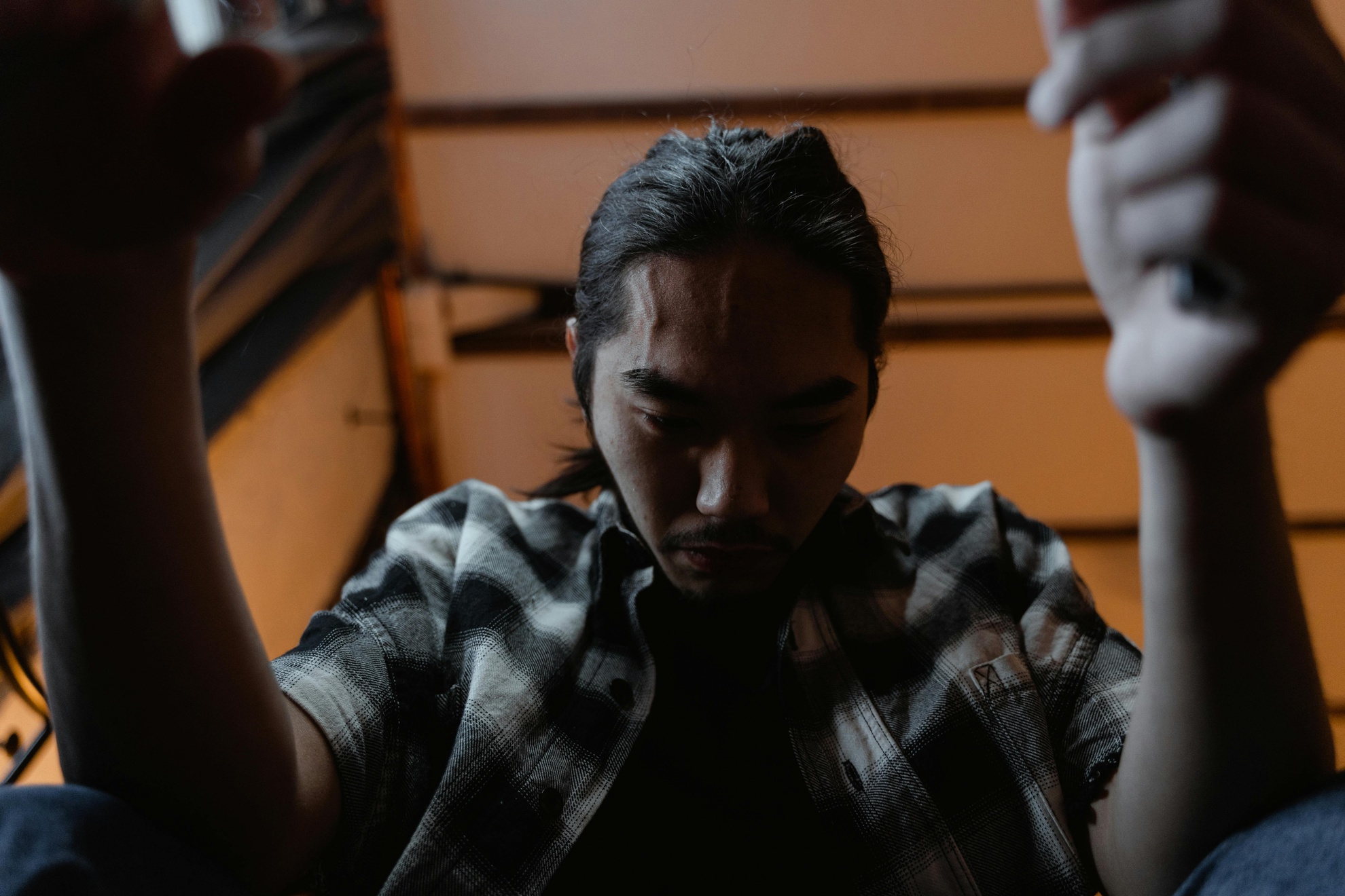 From Renew Hope and Healing: A person with long hair sits indoors, looking down with a serious expression, wearing a plaid shirt. Their hands are raised near the camera, partially out of frame, creating an introspective mood often seen in adult therapy sessions.
