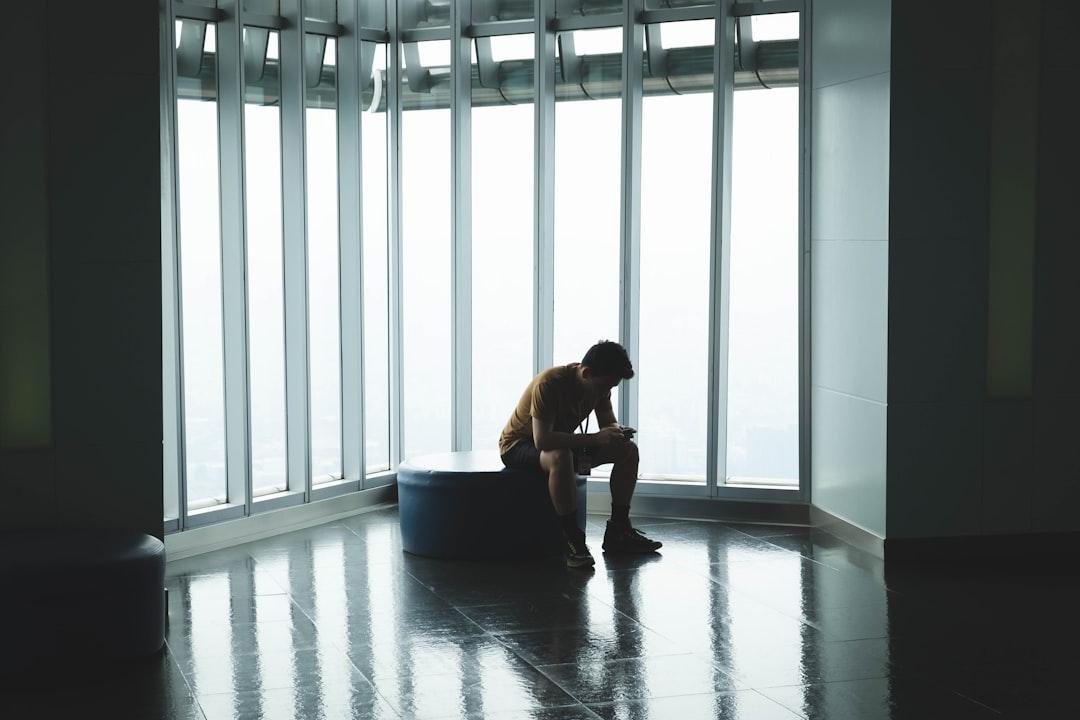 From Renew Hope and Healing: A person sits alone on a round bench by large windows in a high-rise building, reflecting quietly as they look down at a device in their hands—perhaps seeking comfort or support through adult therapy resources.