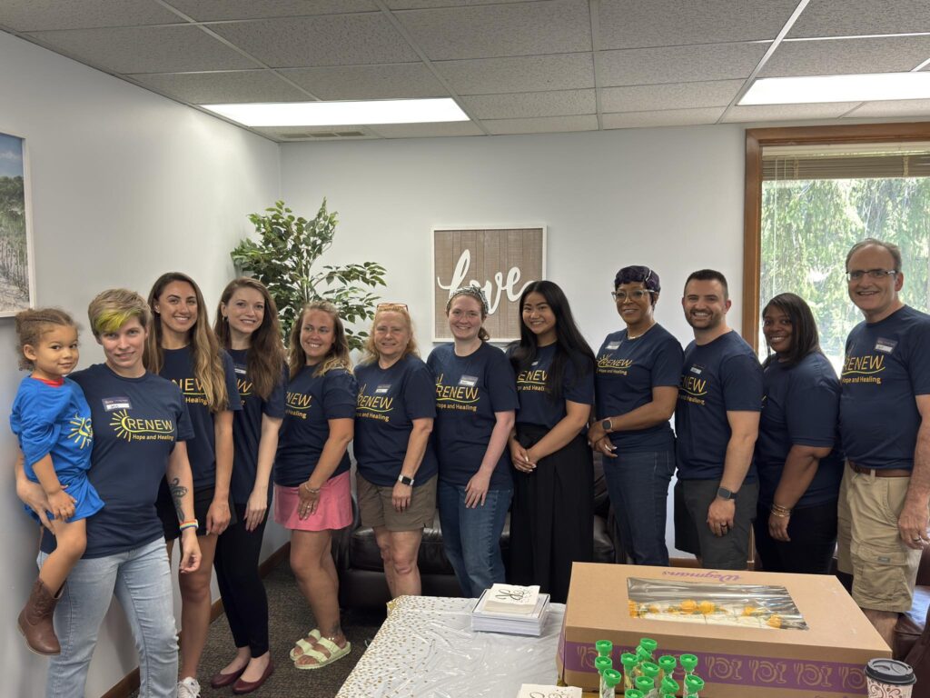 From Renew Hope and Healing: A group of twelve adults and one child stand smiling in an office, all wearing matching navy "RENEW" t-shirts to support youth mental health. A table with refreshments and decorations is in the foreground.