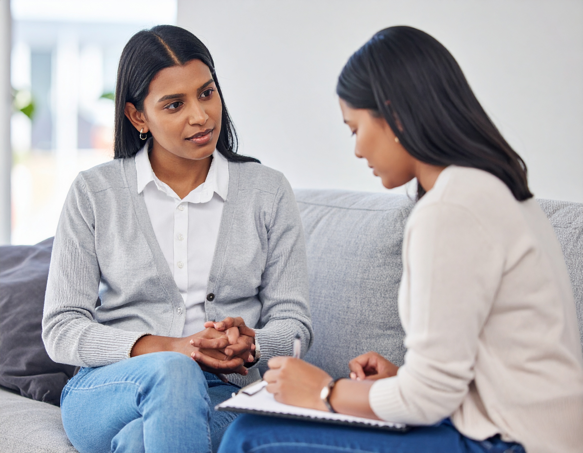 From Renew Hope and Healing: Two women sit on a couch facing each other during a couples counseling session; one listens attentively while the other writes notes on a clipboard.
