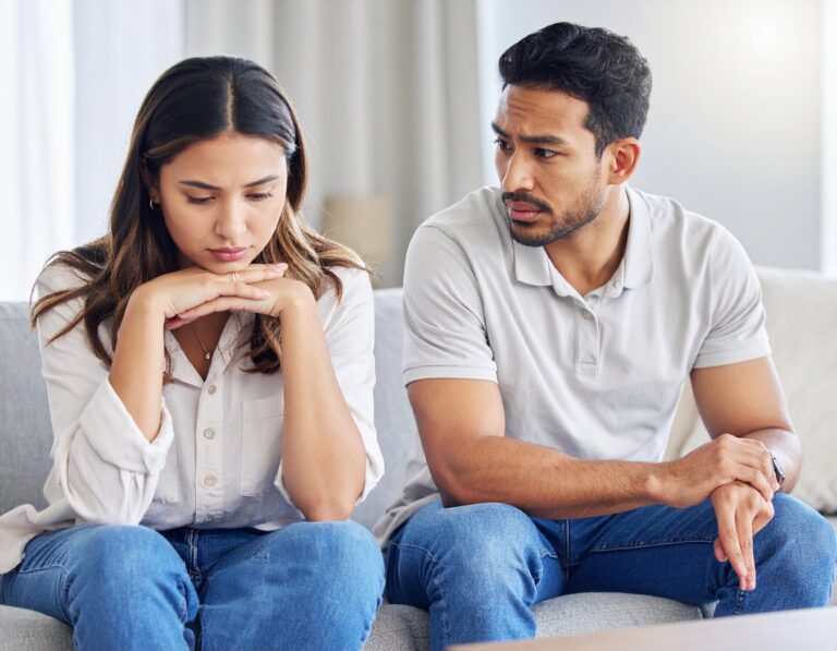 From Renew Hope and Healing: A woman sits on a couch looking down with her hands under her chin, while a man beside her looks at her and gestures with his hand during a couples counseling session.