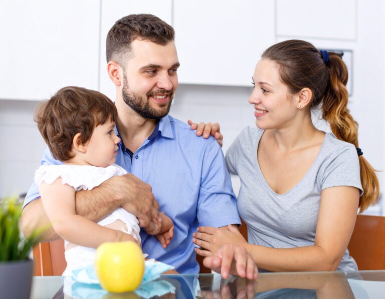 From Renew Hope and Healing: A man holding a toddler sits at a table next to a woman; they are smiling at each other in a bright kitchen setting. An apple is on the table, reflecting the warmth often found after couples counseling sessions.