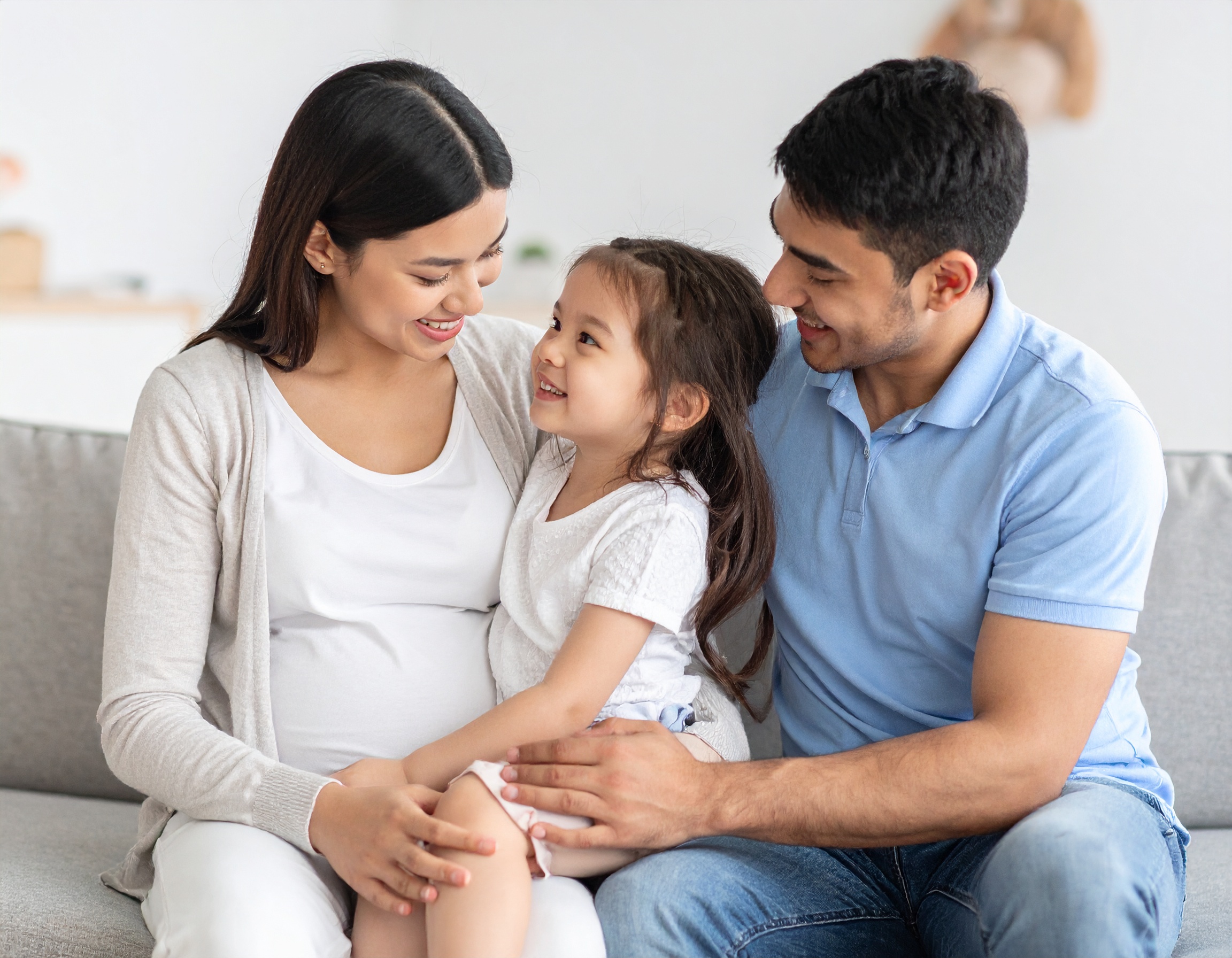 From Renew Hope and Healing: A pregnant woman sits on a couch with a man and a young girl, all smiling and looking at each other in a well-lit living room—capturing the warmth and connection that couples counseling can foster.