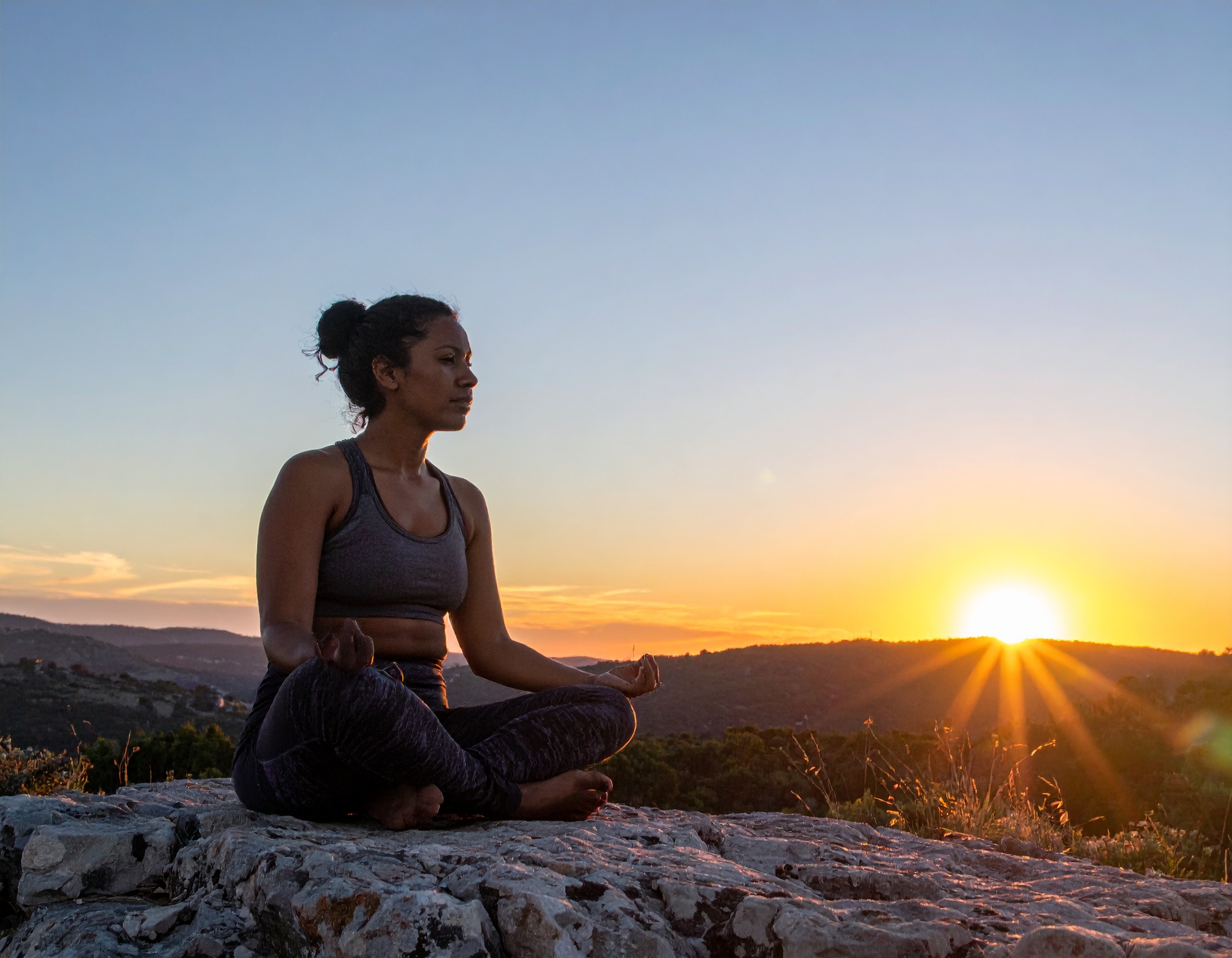 From Renew Hope and Healing: A woman sits cross-legged on a rocky surface, meditating outdoors at sunrise with hills and the sun visible in the background, embracing a peaceful moment often encouraged in adult therapy.