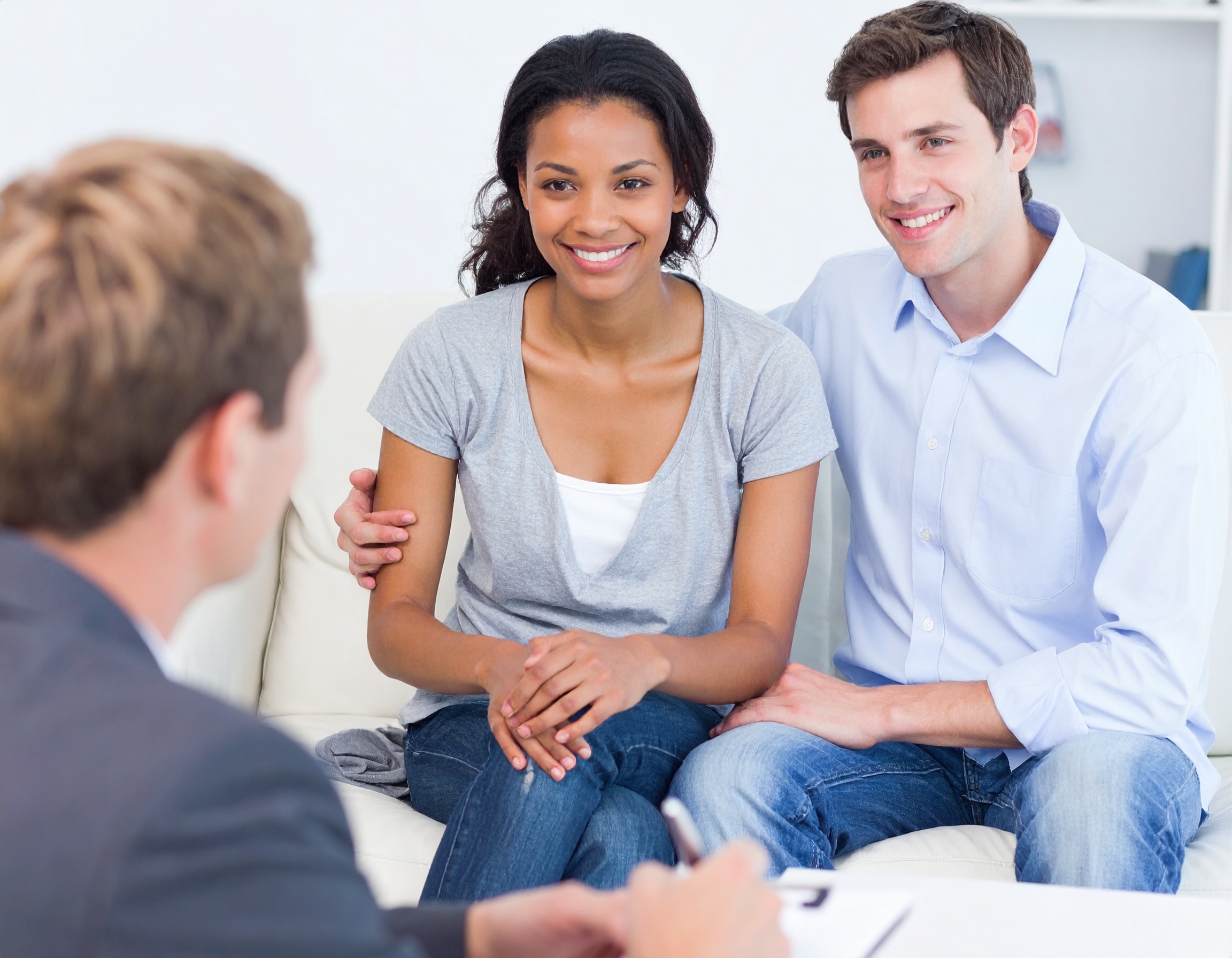 From Renew Hope and Healing: A couple sits on a couch, smiling and listening to a person taking notes across from them during a couples counseling session.