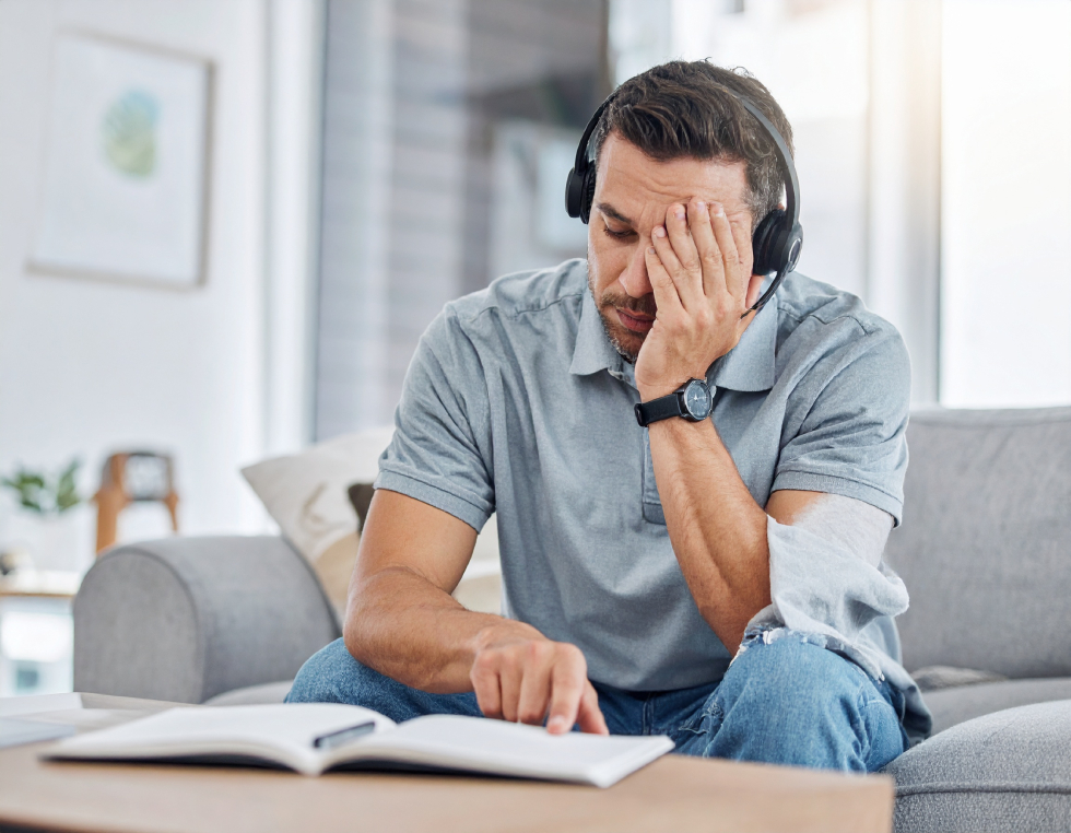 From Renew Hope and Healing: Man sitting on a couch wearing headphones, covering his face with one hand and reading a book with the other, perhaps seeking comfort or exploring anxiety treatment.