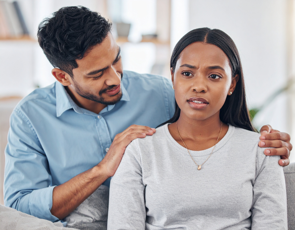 From Renew Hope and Healing: A man sits beside a woman, gently placing his hand on her shoulder in a supportive gesture. The woman looks concerned, suggesting she may benefit from anxiety counseling. Both are indoors and wearing casual clothes.