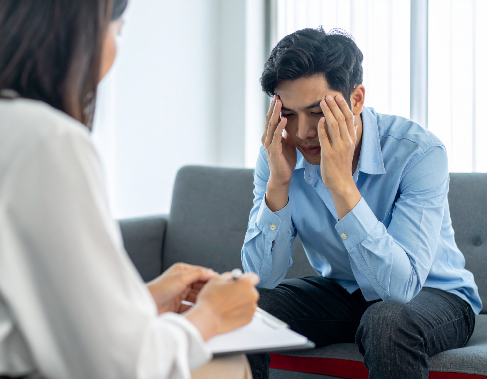 From Renew Hope and Healing: A man sits on a couch holding his head in his hands, appearing stressed, while a woman in the foreground takes notes during an anxiety therapy session.