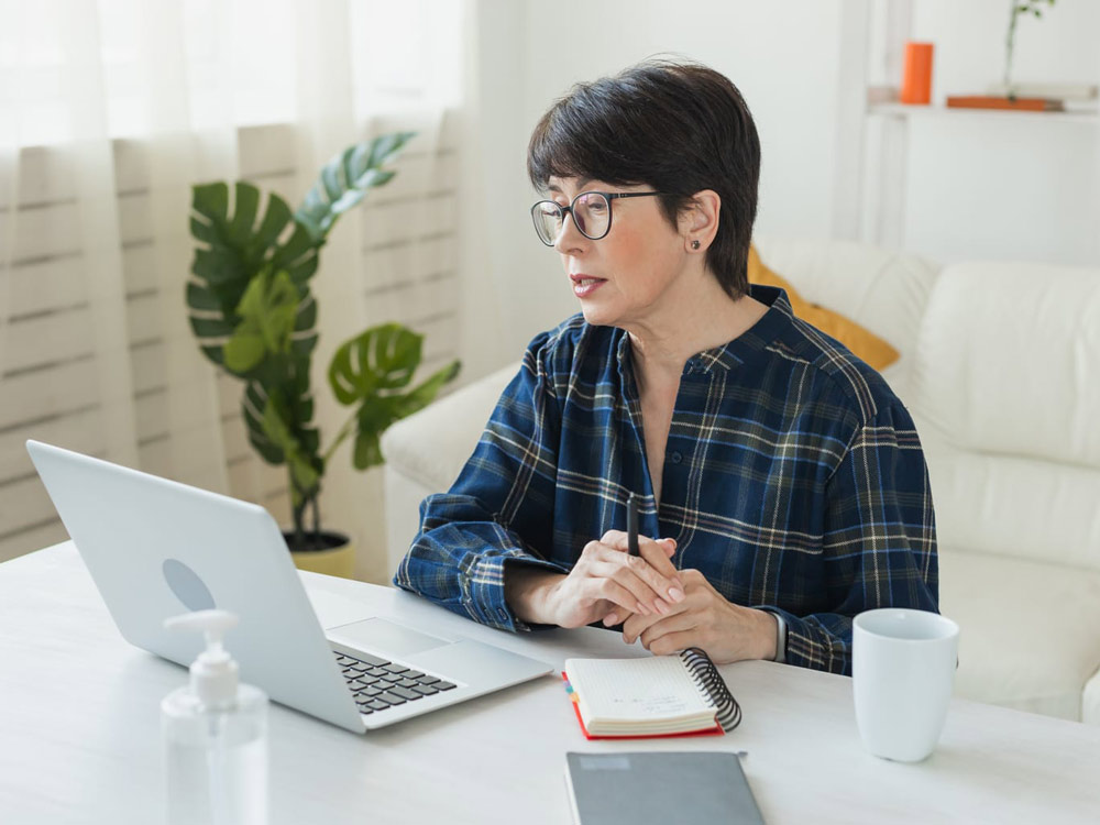 From Renew Hope and Healing: A woman with short hair and glasses sits at a desk in a bright, modern room, looking at her laptop as she researches depression counseling, with a notebook, pen, and coffee mug in front of her.