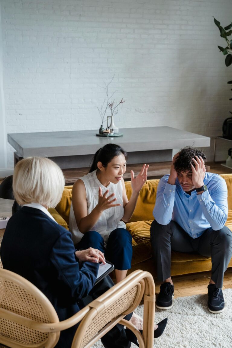 From Renew Hope and Healing: Three people sit indoors on chairs and a sofa; during a couples counseling session, one woman gestures while speaking, a man holds his head in distress, and another woman observes with a notepad.