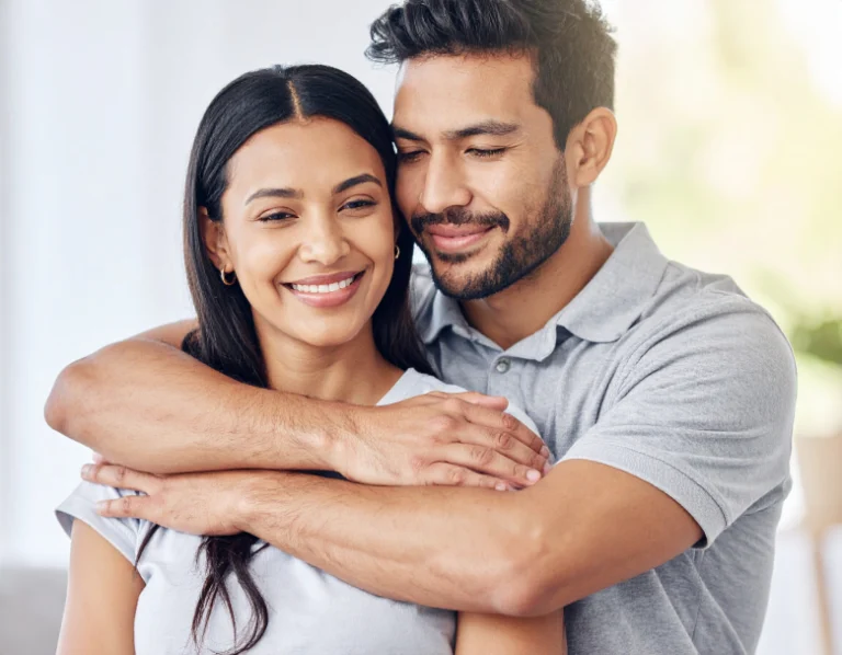 From Renew Hope and Healing: A man smiles while hugging a woman from behind; both are wearing casual clothes and standing indoors, reflecting the warmth and connection often strengthened through couples counseling.