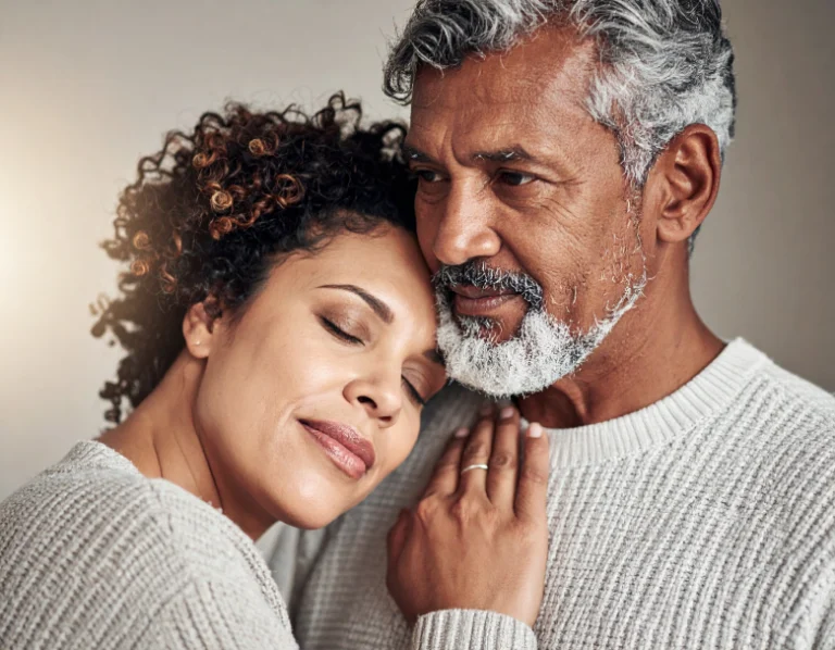 From Renew Hope and Healing: A woman rests her head on a man's chest with her eyes closed, while the man looks forward. Both wear light-colored sweaters and appear calm and content, reflecting the comfort that can come from couples counseling.