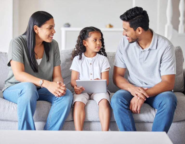 From Renew Hope and Healing: A woman, a young girl holding a tablet, and a man sit together on a gray couch, smiling—capturing a warm moment that reflects the supportive environment of family counseling.