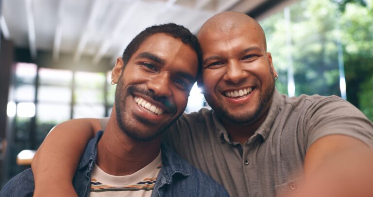 From Renew Hope and Healing: Two men smiling and posing for a selfie indoors, with one man's arm around the other's shoulders—capturing a joyful moment together after a couples counseling session. Natural light streams in from windows in the background.