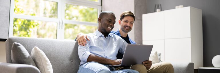 From Renew Hope and Healing: Two men sit together on a couch, smiling and looking at a laptop screen in a bright living room, as they attend an online couples counseling session.