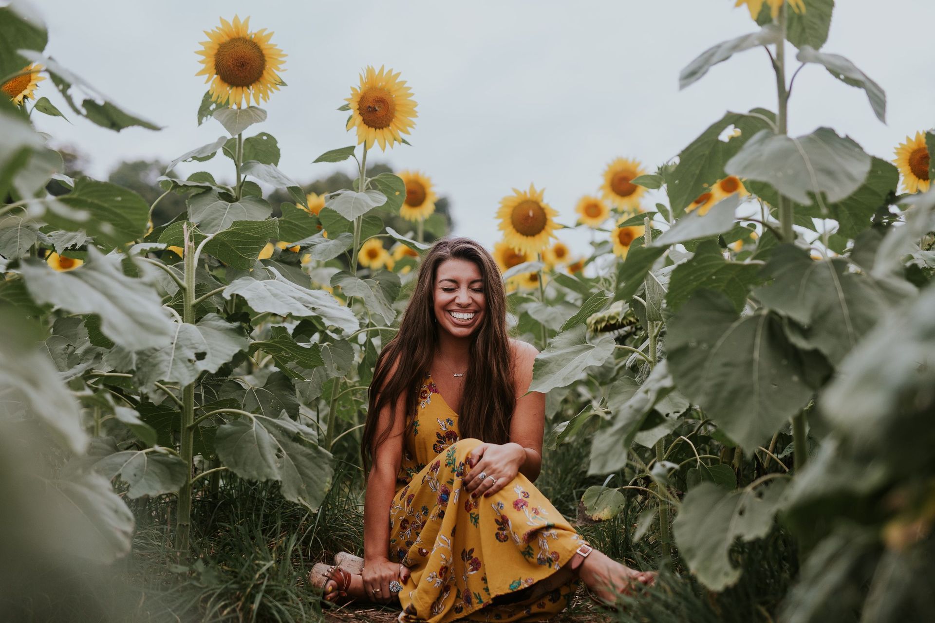 A woman in a yellow floral dress sits smiling in a field of tall sunflowers under a cloudy sky. from Renew Hope and Healing